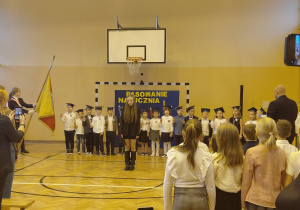 Uczniowie klasy pierwszej stoją w szeregu na salli gimnastycznej, ubrani w stroje galowe i birety na głowach. Na środku stoi uczennica recytująca tekst podczas uroczystości. W tle widoczny jest napis Pasowanie na ucznia. Z przodu stoją pozostali uczniowie i rodzice obserwują występ.